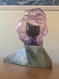 low angle back view. the cat looks very small and soft, the glass pod looks really purple and shiny and blobby and the rock is still angular and geometrical