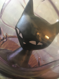 a closeup of the kitty through the glass. it's body is a little conical shape and it's eyes are wide. the face is pointy at the bottom and it has a little white nose. the ears are tall, pointy cones. the glass is reflecting yellow light off od the kitty's forehead. some gray wall and wooden table can be seen behind.