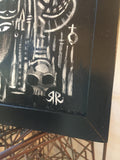 detail shot of the bottom right corner of the painting with Rob reger's signature with a little black circle of space. the top right of that circle aligns with the cheekbone and teeth of a skull. it stands in front of a row of vertical lines 