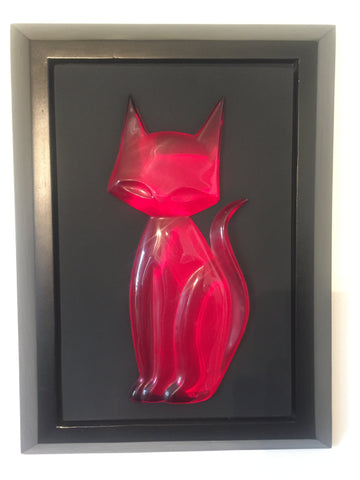 head on photo of a slightly pink-tinted red transparent kitty gem sitting upright and facing left but looking head-on, tail going up in a mild s shape kitty gem is attached to a black mat and set in a black frame.
