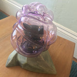 top-front view of the sculpture, the kitty's tail curls slightly toward the back and the right eye is visible. the left eye is obscured by the warped glass. it sits on a wooden table in front of a gray wall. a bit of blue carpet is visible below.