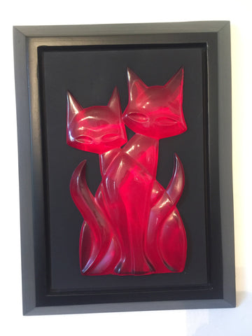 head on photo of 2 transparent red cads sitting one in front of the other facing the viewer. they form a single shape that appears to have 2 tails, one on either side. this sits on a black mat set in a black frame